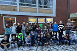 Das Foto zeigt die Klasse 6a mit einem Schneemann auf dem Pausenhof des TGG und ihrem grünen Klassenmaskottchen (Dezember 2023).